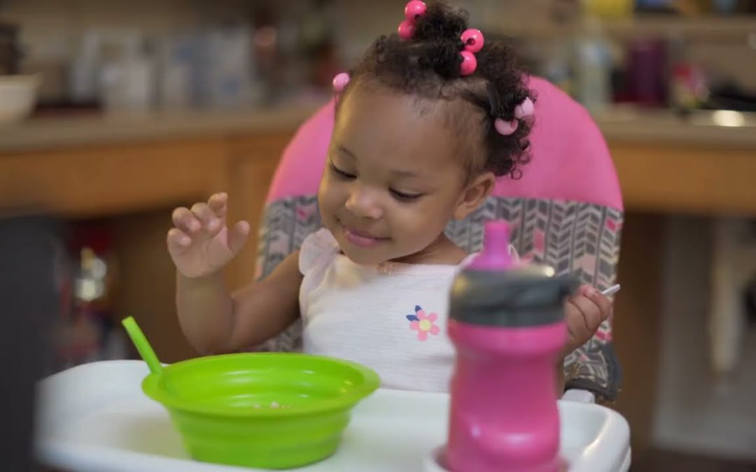 A little girl sitting in her high chair, smiling down at her bowl of food.