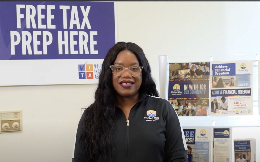 A woman standing, smiling in front of the Volunteer Income Tax Assistance sign